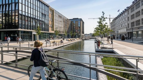 Blick auf das neu gestaltete Holstenfleet in Kiel mit Wasserlauf, Fußgängerbereich und Sitzmöglichkeiten; eine Radfahrerin überquert im Vordergrund eine Brücke.