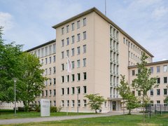 Sechsgeschossiger Verwaltungsbau in hellem Putz; regelmäßige Fensterreihen, Rasen und Bäume davor; Eingang mit Flagge und Schild.