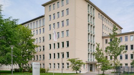 Sechsgeschossiger Verwaltungsbau in hellem Putz; regelmäßige Fensterreihen, Rasen und Bäume davor; Eingang mit Flagge und Schild.