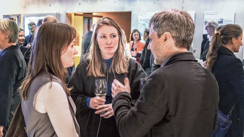 „Zwei Frauen und ein Mann unterhalten sich bei einer gut besuchten Veranstaltung. Eine der Frauen hält ein Weinglas in den Händen.“