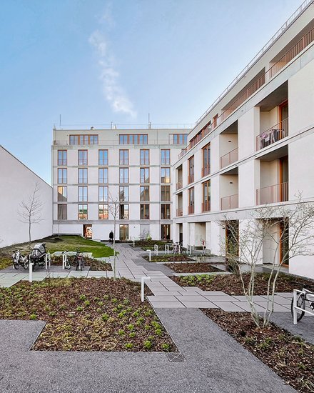 "Innenhof eines modernen Wohnkomplexes mit zwei mehrstöckigen, hellen Gebäuden. Der Hof ist gepflastert mit grauen Steinplatten und begrünten Bereichen mit Mulch und jungen Bäumen. Rechts ein weißes Gebäude mit mehreren Balkonen und großen Fenstern. Links ein Gebäude mit Holzdetails und Metallgeländer. Ein Fahrradständer mit Fahrrädern steht links. Der Himmel ist blau mit wenigen Wolken. Die Anlage wirkt modern, gepflegt und verkehrsfrei."