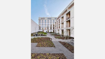 "Innenhof eines modernen Wohnkomplexes mit zwei mehrstöckigen, hellen Gebäuden. Der Hof ist gepflastert mit grauen Steinplatten und begrünten Bereichen mit Mulch und jungen Bäumen. Rechts ein weißes Gebäude mit mehreren Balkonen und großen Fenstern. Links ein Gebäude mit Holzdetails und Metallgeländer. Ein Fahrradständer mit Fahrrädern steht links. Der Himmel ist blau mit wenigen Wolken. Die Anlage wirkt modern, gepflegt und verkehrsfrei."