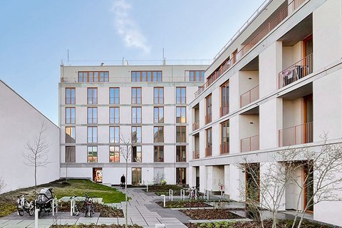 "Innenhof eines modernen Wohnkomplexes mit zwei mehrstöckigen, hellen Gebäuden. Der Hof ist gepflastert mit grauen Steinplatten und begrünten Bereichen mit Mulch und jungen Bäumen. Rechts ein weißes Gebäude mit mehreren Balkonen und großen Fenstern. Links ein Gebäude mit Holzdetails und Metallgeländer. Ein Fahrradständer mit Fahrrädern steht links. Der Himmel ist blau mit wenigen Wolken. Die Anlage wirkt modern, gepflegt und verkehrsfrei."