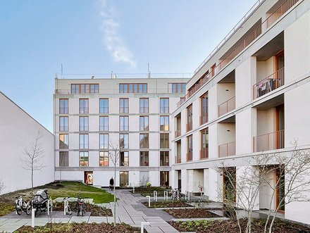 "Innenhof eines modernen Wohnkomplexes mit zwei mehrstöckigen, hellen Gebäuden. Der Hof ist gepflastert mit grauen Steinplatten und begrünten Bereichen mit Mulch und jungen Bäumen. Rechts ein weißes Gebäude mit mehreren Balkonen und großen Fenstern. Links ein Gebäude mit Holzdetails und Metallgeländer. Ein Fahrradständer mit Fahrrädern steht links. Der Himmel ist blau mit wenigen Wolken. Die Anlage wirkt modern, gepflegt und verkehrsfrei."