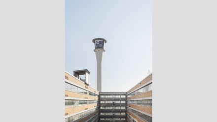Innenhof mit markantem Turm  Ein Blick nach oben in einen rechteckigen Innenhof. Die Fassaden bestehen aus Backstein und horizontalen Fensterbändern. In der Mitte ragt ein hoher, schlanker Turm mit einer technischen Aufbauten an der Spitze in den Himmel. Der Innenhof bringt Licht und Luft in den Gebäudekomplex. Der Turm dient als technisches oder infrastrukturelles Element und ist ein prägnantes Erkennungszeichen des Ortes.
