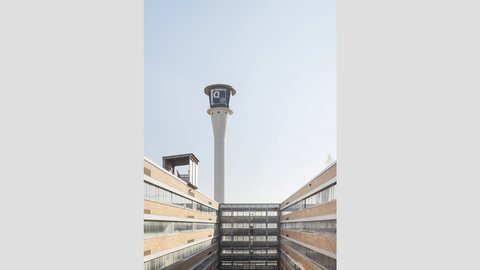 Innenhof mit markantem Turm  Ein Blick nach oben in einen rechteckigen Innenhof. Die Fassaden bestehen aus Backstein und horizontalen Fensterbändern. In der Mitte ragt ein hoher, schlanker Turm mit einer technischen Aufbauten an der Spitze in den Himmel. Der Innenhof bringt Licht und Luft in den Gebäudekomplex. Der Turm dient als technisches oder infrastrukturelles Element und ist ein prägnantes Erkennungszeichen des Ortes.