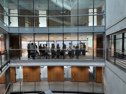 "Blick in ein modernes Bürogebäude mit zwei übereinander liegenden Galerien. Die obere Galerie zeigt eine Gruppe von etwa 20 Personen, die in einem hellen Konferenzraum mit großen Fenstern stehen. Weiße Decken, graue Betonwände und Holzverkleidungen prägen die Architektur."