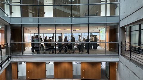 "Blick in ein modernes Bürogebäude mit zwei übereinander liegenden Galerien. Die obere Galerie zeigt eine Gruppe von etwa 20 Personen, die in einem hellen Konferenzraum mit großen Fenstern stehen. Weiße Decken, graue Betonwände und Holzverkleidungen prägen die Architektur."