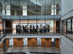 "Blick in ein modernes Bürogebäude mit zwei übereinander liegenden Galerien. Die obere Galerie zeigt eine Gruppe von etwa 20 Personen, die in einem hellen Konferenzraum mit großen Fenstern stehen. Weiße Decken, graue Betonwände und Holzverkleidungen prägen die Architektur."
