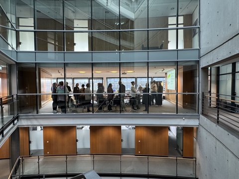 "Blick in ein modernes Bürogebäude mit zwei übereinander liegenden Galerien. Die obere Galerie zeigt eine Gruppe von etwa 20 Personen, die in einem hellen Konferenzraum mit großen Fenstern stehen. Weiße Decken, graue Betonwände und Holzverkleidungen prägen die Architektur."