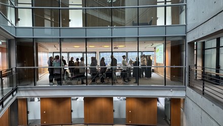 "Blick in ein modernes Bürogebäude mit zwei übereinander liegenden Galerien. Die obere Galerie zeigt eine Gruppe von etwa 20 Personen, die in einem hellen Konferenzraum mit großen Fenstern stehen. Weiße Decken, graue Betonwände und Holzverkleidungen prägen die Architektur."