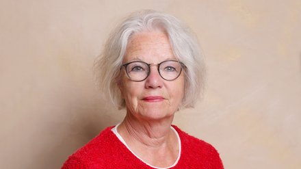 "Porträt einer älteren Frau mit weißem, schulterlangem Haar und Brille mit dunklem Rand. Sie trägt ein rotes Fleece-Shirt mit weißem Ausschnitt und einen kleinen roten Knopf-Anstecker. Hintergrund in Beige-Creme-Tönen."