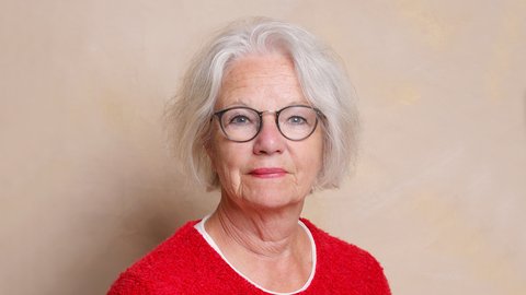 "Porträt einer älteren Frau mit weißem, schulterlangem Haar und Brille mit dunklem Rand. Sie trägt ein rotes Fleece-Shirt mit weißem Ausschnitt und einen kleinen roten Knopf-Anstecker. Hintergrund in Beige-Creme-Tönen."