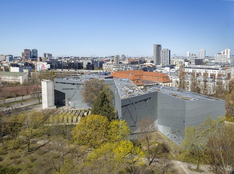 Luftaufnahme eines modernen, grauen Gebäudes mit schrägen Wänden umgeben von Bäumen und einer Stadt im Hintergrund