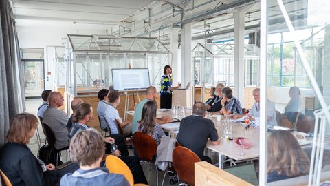 „Eine Frau hält eine Präsentation vor einer sitzenden Gruppe von Zuhörern in einem hellen, modern gestalteten Konferenzraum im Industrie-Stil.“