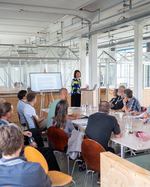 „Eine Frau hält eine Präsentation vor einer sitzenden Gruppe von Zuhörern in einem hellen, modern gestalteten Konferenzraum im Industrie-Stil.“