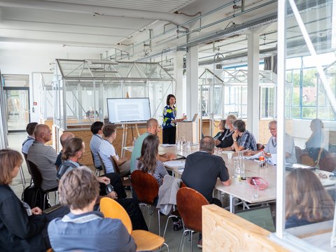 „Eine Frau hält eine Präsentation vor einer sitzenden Gruppe von Zuhörern in einem hellen, modern gestalteten Konferenzraum im Industrie-Stil.“