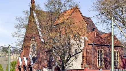 Backsteinkirche mit Turm und spitzem Dach, umgeben von Bäumen an einer Straße.