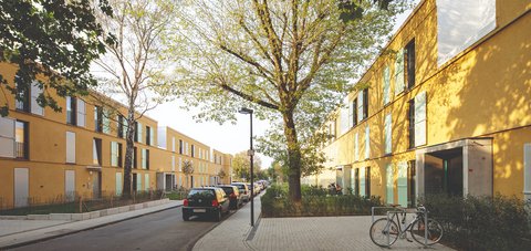 Ruhige Wohnstraße mit dichtem Baumdach; beidseitig zweigeschossige, ockerfarbene Reihenhäuser mit mintgrünen Fensterläden; geparkte Autos und ein Fahrradständer auf breiten Gehwegen im Schatten.