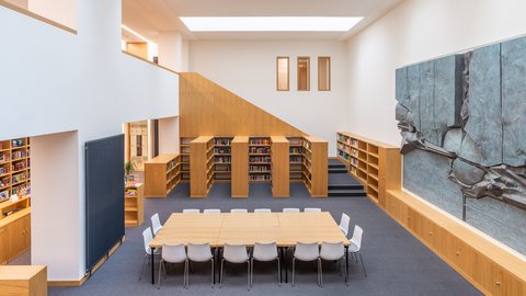 Moderner, heller Leseraum mit großem Holztisch, weißen Stühlen, Bücherregalen und einer großen Wandskulptur