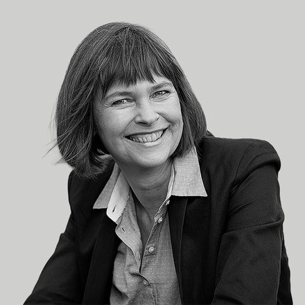 Schwarzweiß‑Porträt einer lächelnden Frau in Halbnahaufnahme; kinnlanges Haar mit Pony, dunkel jacket und helles Hemd, leicht zur Seite geneigt, offener, freundlicher Ausdruck vor einfarbigem Hintergrund.