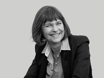 Schwarzweiß‑Porträt einer lächelnden Frau in Halbnahaufnahme; kinnlanges Haar mit Pony, dunkel jacket und helles Hemd, leicht zur Seite geneigt, offener, freundlicher Ausdruck vor einfarbigem Hintergrund.