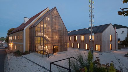 "Moderne Kirche mit beleuchteter, heller Fassade und Holzlamellenelement. Links ein weißes Nebengebäude, rechts ein Gebäude mit vertikaler Holzstruktur. Eine beleuchtete Säule mit religiösen Symbolen steht vor dem Gebäude. Abendliche Aufnahme mit warmen Lichtern in den Fenstern."