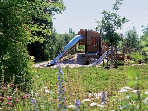 Naturnaher Spielplatz mit Holzspielhaus, Rutschen und Kletterelementen in einer grünen Umgebung.