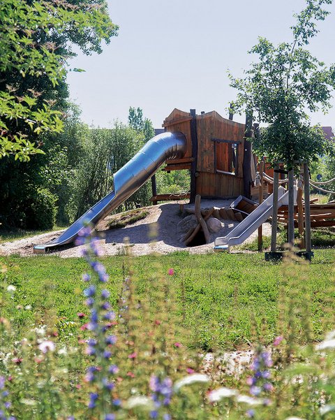 Naturnaher Spielplatz mit Holzspielhaus, Rutschen und Kletterelementen in einer grünen Umgebung.