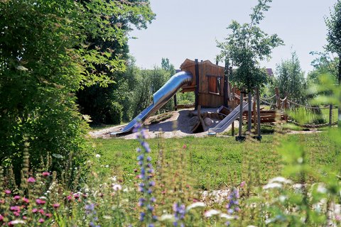 Naturnaher Spielplatz mit Holzspielhaus, Rutschen und Kletterelementen in einer grünen Umgebung.