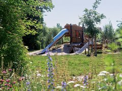 Naturnaher Spielplatz mit Holzspielhaus, Rutschen und Kletterelementen in einer grünen Umgebung.