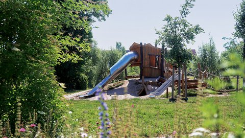 Naturnaher Spielplatz mit Holzspielhaus, Rutschen und Kletterelementen in einer grünen Umgebung.