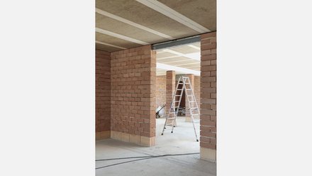 Rohbau-Innenraum aus roten Ziegeln mit Stützen und Öffnungen; Leiter und Werkzeuge, Decke mit sichtbaren Holzträgern.