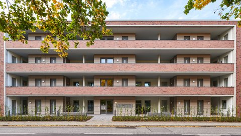 Symmetrische Frontalansicht eines mehrstöckigen Wohnhauses. Die Fassade wird durch lange, horizontale Balkonbänder aus rotem Backstein gegliedert. Hinter den Balkonen sind helle, beige Wandelemente sowie graue Fenster und Türen zu sehen. Im Vordergrund verläuft ein Gehweg mit einer niedrigen Hecke; links oben ragen herbstlich gefärbte Baumblätter ins Bild.