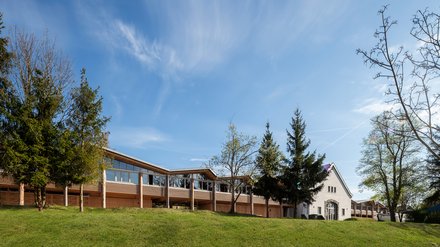 Fernansicht: langes Holzgebäude auf Stützen neben weißem Bestandsbau, umgeben von Wiese und Bäumen.