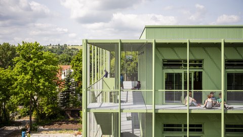 Grün verkleidetes Schulgebäude mit außenliegender Treppe und galerieartigen Gängen hinter Metallnetzen. Kinder sitzen und unterhalten sich auf dem oberen Gang; links rahmen Bäume und ein bewölkter Himmel die Szene.