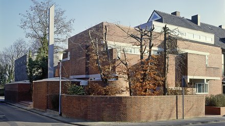 "Modernes Backsteingebäude mit Satteldach an einer Straßenecke. Rote Backsteinmauern, Gartenmauer aus Backstein, kahle Bäume. Ein dunkler Quader-Anbau mit verglaster Fassade auf der linken Seite. Klarer Himmel."