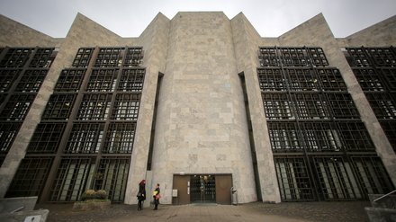 Eine monumentale, symmetrisch aufgebaute Gebäudefassade aus hellem Stein mit stark gegliederten Wandflächen und tiefen Fenstergittern dominiert den Stadtraum, der zentrale Eingang ist zurückgesetzt und wird von wenigen Personen im Maßstab erfahrbar gemacht, wodurch das Bild ein bedeutendes Verwaltungs- oder Kulturbauwerk zeigt und für die Auseinandersetzung mit historischer Architektur, Denkmalwert und zeitgenössischer Nutzung steht.