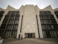 Eine monumentale, symmetrisch aufgebaute Gebäudefassade aus hellem Stein mit stark gegliederten Wandflächen und tiefen Fenstergittern dominiert den Stadtraum, der zentrale Eingang ist zurückgesetzt und wird von wenigen Personen im Maßstab erfahrbar gemacht, wodurch das Bild ein bedeutendes Verwaltungs- oder Kulturbauwerk zeigt und für die Auseinandersetzung mit historischer Architektur, Denkmalwert und zeitgenössischer Nutzung steht.