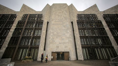 Eine monumentale, symmetrisch aufgebaute Gebäudefassade aus hellem Stein mit stark gegliederten Wandflächen und tiefen Fenstergittern dominiert den Stadtraum, der zentrale Eingang ist zurückgesetzt und wird von wenigen Personen im Maßstab erfahrbar gemacht, wodurch das Bild ein bedeutendes Verwaltungs- oder Kulturbauwerk zeigt und für die Auseinandersetzung mit historischer Architektur, Denkmalwert und zeitgenössischer Nutzung steht.