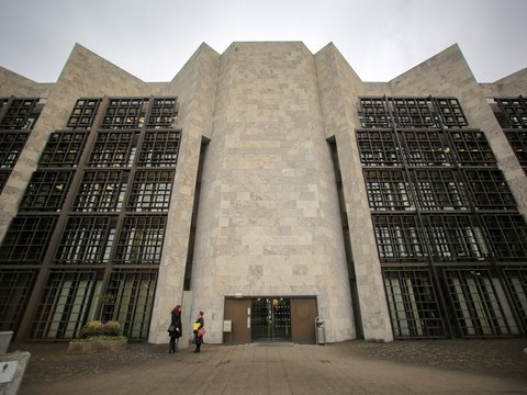 Eine monumentale, symmetrisch aufgebaute Gebäudefassade aus hellem Stein mit stark gegliederten Wandflächen und tiefen Fenstergittern dominiert den Stadtraum, der zentrale Eingang ist zurückgesetzt und wird von wenigen Personen im Maßstab erfahrbar gemacht, wodurch das Bild ein bedeutendes Verwaltungs- oder Kulturbauwerk zeigt und für die Auseinandersetzung mit historischer Architektur, Denkmalwert und zeitgenössischer Nutzung steht.
