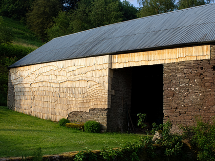 Ländliche Scheune mit Wellblechdach; Wände teils aus Bruchstein, teils mit hellen Holzschindeln verkleidet; davor eine grüne Wiese, dahinter Bäume.