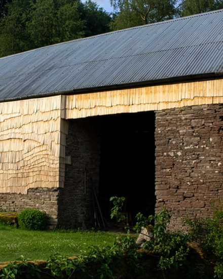 Ländliche Scheune mit Wellblechdach; Wände teils aus Bruchstein, teils mit hellen Holzschindeln verkleidet; davor eine grüne Wiese, dahinter Bäume.