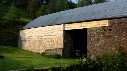 Ländliche Scheune mit Wellblechdach; Wände teils aus Bruchstein, teils mit hellen Holzschindeln verkleidet; davor eine grüne Wiese, dahinter Bäume.