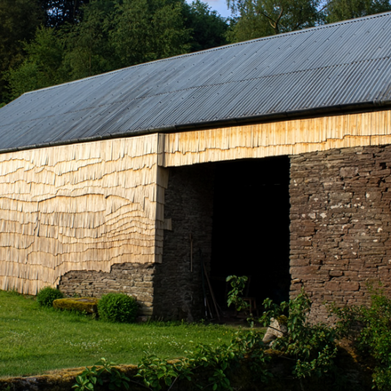 Ländliche Scheune mit Wellblechdach; Wände teils aus Bruchstein, teils mit hellen Holzschindeln verkleidet; davor eine grüne Wiese, dahinter Bäume.