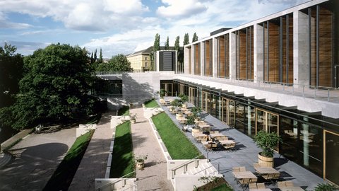 Modernes Gebäude mit Betonfassade und großflächigen Fenstern,  umgeben von gepflegtem Innenhof. Gepflasterte und grasige Bereiche  mit Sitzmöbeln, Bäumen in Kübeln, Treppen führen zu erhöhten  Bereichen, historische Gebäude im Hintergrund.