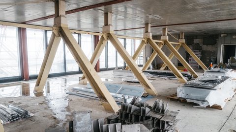 Rohbau-Etage mit gelben, schrägen Stahlstützen entlang einer verglasten Fassade; nasser Betonboden, gestapelte Metallteile und Paletten mit verpackten Bauteilen.