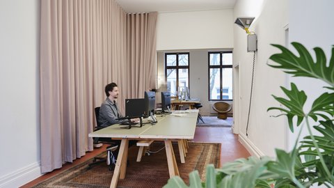 Mann sitzt in Büro an einem Schreibtisch. Roter Boden mit Teppich und grüne Pflanze im Vordergrund