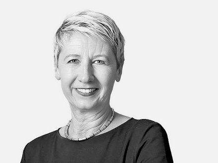 Schwarz-Weiß-Porträt einer Frau mit kurzen, hellgrauen Haaren, die direkt in die Kamera lächelt. Sie trägt ein schwarzes Shirt und eine silberfarbene Halskette. Der Hintergrund ist weiß.