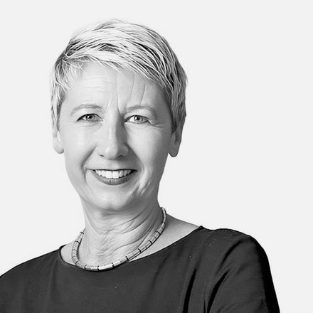 Schwarz-Weiß-Porträt einer Frau mit kurzen, hellgrauen Haaren, die direkt in die Kamera lächelt. Sie trägt ein schwarzes Shirt und eine silberfarbene Halskette. Der Hintergrund ist weiß.